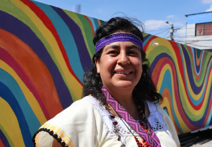 A person wearing traditional clothing and a purple headband stands in front of a colourful mural with wavy patterns.